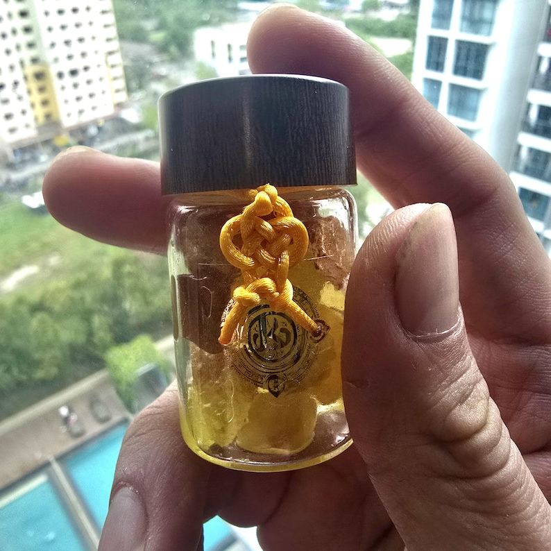 Puede incluir: Una peque&ntilde;a botella de vidrio con tapa de madera oscura, llena de contenido amarillo. Un cord&oacute;n amarillo anudado est&aacute; atado alrededor de la botella, con un emblema dorado. La botella est&aacute; sostenida en la mano de una persona.