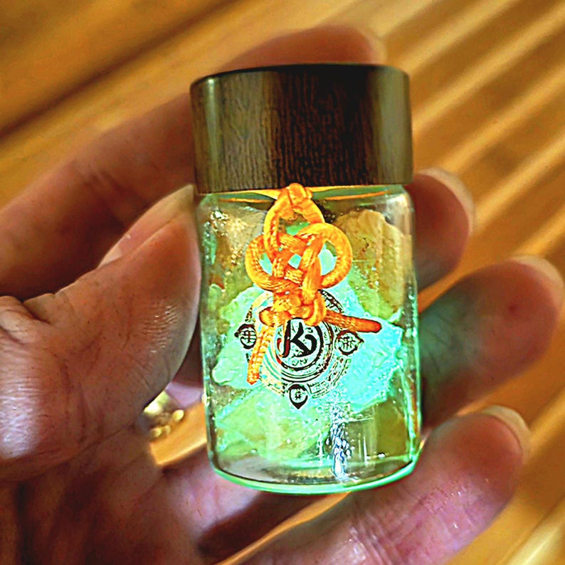 Puede incluir: Una peque&ntilde;a botella de vidrio con tapa de madera oscura, llena de un l&iacute;quido verde claro y objetos verdes y amarillos p&aacute;lidos. La botella est&aacute; adornada con un nudo naranja decorativo y una etiqueta con un dise&ntilde;o estilizado "K".