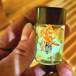 Puede incluir: Una peque&ntilde;a botella de vidrio con tapa de madera oscura, llena de l&iacute;quido amarillo. La botella est&aacute; adornada con un cord&oacute;n anaranjado anudado y una etiqueta con un logotipo. La botella se sostiene en una mano.