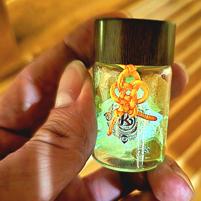 Puede incluir: Una peque&ntilde;a botella de vidrio con tapa de madera oscura, llena de l&iacute;quido amarillo. La botella est&aacute; adornada con un cord&oacute;n anaranjado anudado y una etiqueta con un logotipo. La botella se sostiene en una mano.