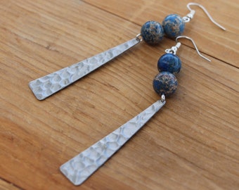 May include: A pair of dangle earrings featuring hammered silver-tone metal and blue and brown jasper beads. The earrings have a long, rectangular shape with a textured surface and are suspended from silver-tone ear wires. The beads add a pop of color.