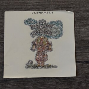 May include: A square, off-white card featuring a glittery illustration of a figure with a mushroom cloud above. The figure is composed of various colors, with silver and blue glitter dominating the cloud. The text "D3219M-R41438" is printed at the top.