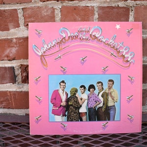 May include: A vintage vinyl record album cover for "Johnny and the RocKet88s" in pink with a blue rectangle featuring a band photo. The album art includes the band's name in a stylized font, small airplane graphics, and a star.