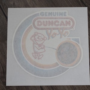 May include: A vintage sticker featuring the text "GENUINE DUNCAN Yo-Yo" in a retro design. The sticker has a circular design with a cartoon figure playing with a yo-yo. The color scheme includes red, blue, and orange.