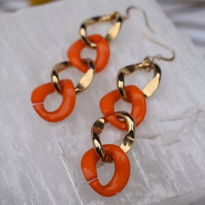 May include: A pair of gold-tone chain link earrings with bright orange enamel accents. The earrings feature a series of interlocking links, with the orange links alternating with gold-colored links. The earrings are displayed on a white, textured surface.
