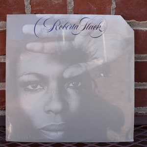 May include: A vinyl record album cover featuring a black and white image of a woman's face with a hand partially covering it. The album title "Roberta Flack" is written in elegant script across the top. The background is a red brick wall.