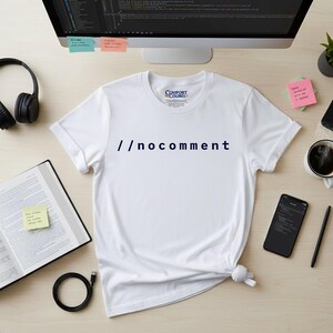 May include: White t-shirt with the text "//nocomment" in navy blue. The shirt is on a wooden desk with a computer monitor, headphones, a book, a phone, and a cup of coffee.