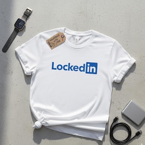 May include: White t-shirt with rolled sleeves displaying the blue text "LockedIn" and a blue square. A small tag reads "Handmade with Code". A black watch, a silver device, and a black cable are also visible.