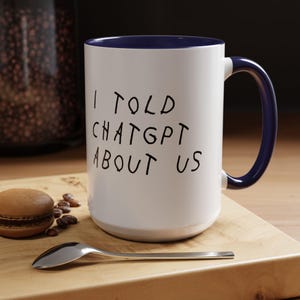 May include: White ceramic mug with a navy blue interior and handle. The mug features the text "I TOLD CHATGPT ABOUT US" in black, handwritten-style lettering. A macaron, coffee beans, and a spoon are on a wooden surface.