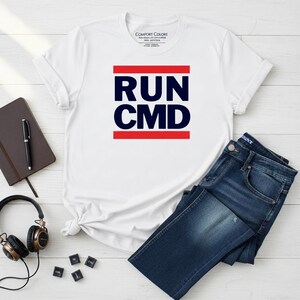 Peut inclure: T-shirt blanc avec l'inscription "RUN CMD" en bleu marine, encadrée de lignes rouges. Le t-shirt est associé à un jean bleu. Un carnet, un stylo et un casque sont également visibles.