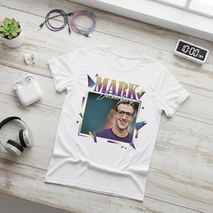 May include: White t-shirt featuring a portrait of Mark Zuckerberg with the name "MARK" in a retro font. The shirt has a colorful geometric design and is surrounded by accessories like headphones and earbuds.