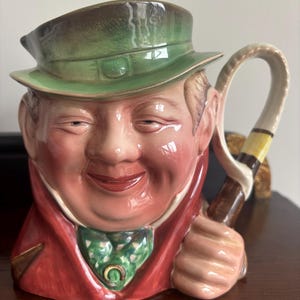 Könnte beinhalten: Ein Keramikkrug in Form eines lächelnden Mannes mit grünem Hut und rotem Mantel. Der Griff ist eine gebogene Peitsche mit gelben und braunen Streifen. Das Gesicht des Mannes ist detailliert mit rosigen Wangen und einer grünen gemusterten Krawatte.