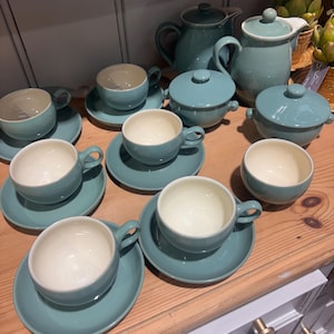 Puede incluir: Colección de tazas de té, platillos, teteras y cuencos cubiertos de cerámica color turquesa, dispuestos sobre una superficie de madera. Las tazas tienen interior blanco. El juego incluye varios tamaños y formas.