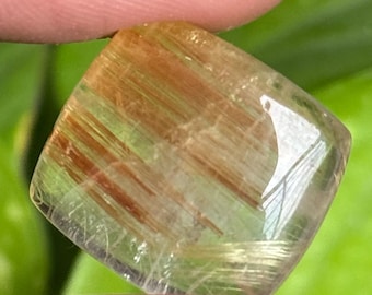 以下が含まれることがあります： 金茶色のインクルージョンが入った、透明な正方形にカットされた宝石。宝石は、緑の葉を背景に、2本の指で持たれています。石は研磨された反射面を持ち、その自然な模様と色を示しています。