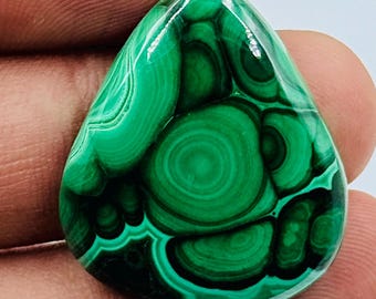 Puede incluir: Una piedra preciosa de malaquita pulida en forma de lágrima que presenta bandas concéntricas vibrantes de verde bosque profundo y verde esmeralda más claro. La piedra tiene una superficie lisa y reflectante que resalta los patrones naturales.