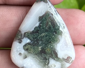 Puede incluir: Una gema en forma de lágrima con una base blanca e intrincadas inclusiones verdes y marrones. La piedra pulida se sostiene en una mano, mostrando sus patrones y texturas naturales. El diseño único de la gema la convierte en una hermosa pieza para joyería o colección.
