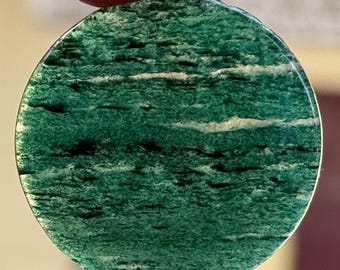 Puede incluir: Una piedra redonda verde con vetas blancas y negras. La piedra se sostiene entre dos dedos. La superficie pulida de la piedra le da un aspecto natural y orgánico. El color es un verde intenso y rico.