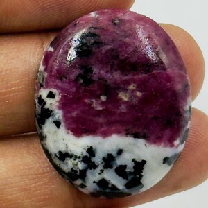 May include: An oval-shaped polished gemstone featuring a vibrant magenta-pink section, a contrasting white area, and scattered black mineral inclusions throughout the surface. The stone has a smooth, glossy finish and a rounded, cabochon-style shape.