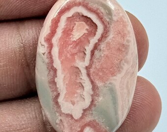 Puede incluir: Un cabujón de rodocrosita de forma ovalada que presenta bandas concéntricas de color rosa suave, blanco crema y gris apagado. La piedra pulida muestra un patrón mineral natural en capas con un acabado de superficie liso y brillante.