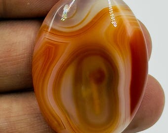 May include: An oval-shaped cabochon gemstone featuring concentric bands of translucent orange, deep red, and creamy white colors. The polished stone displays a smooth, glossy surface with natural swirling patterns and light reflections.