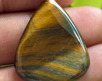 May include: A polished, triangular tiger's eye gemstone. The stone displays bands of golden brown, yellow, and gray hues. The gemstone is held in a hand, with green foliage in the background.