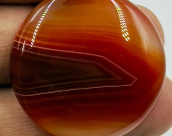 May include: A round, polished carnelian gemstone cabochon featuring vibrant bands of deep orange, red, and dark brown hues. The smooth, glossy surface displays a distinct chevron-like pattern within the stone's natural translucent layers.