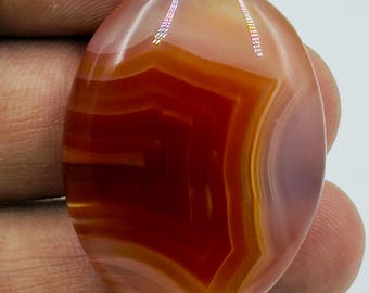 May include: An oval-shaped cabochon gemstone featuring translucent layers of deep orange, reddish-brown, and pale cream colors. The stone displays a distinct banded pattern with a central angular design, polished to a smooth, glossy finish.