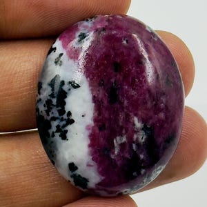 Pode incluir: Uma pedra preciosa polida de formato oval com uma seção rosa magenta vibrante contrastada por uma área branca salpicada de inclusões pretas escuras. A pedra tem uma superfície lisa e brilhante com um corte cabochon arredondado e padrões naturais.