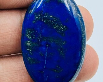 Puede incluir: Una piedra preciosa de lapislázuli de forma ovalada con una superficie pulida y lisa. La piedra presenta un color base azul profundo y vibrante con inclusiones oscuras visibles y sutiles motas doradas dispersas por toda la superficie.