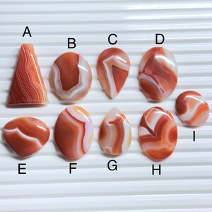 May include: A collection of nine polished, smooth, orange and white banded agate cabochons labeled A through I. The stones feature various shapes including triangles, ovals, and teardrops, each displaying unique natural swirling patterns and layers.