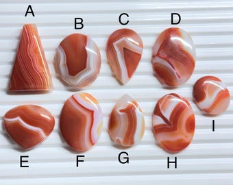 May include: A collection of nine polished, smooth, orange and white banded agate cabochons labeled A through I. The stones feature various shapes including triangles, ovals, and teardrops, each displaying unique natural swirling patterns and layers.