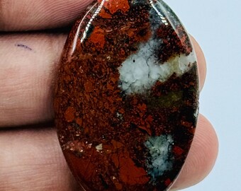 Puede incluir: Una piedra preciosa pulida de forma ovalada que presenta una base de color rojo parduzco profundo con prominentes parches moteados de inclusiones blancas opacas y gris oscuro en toda la superficie lisa y reflectante.
