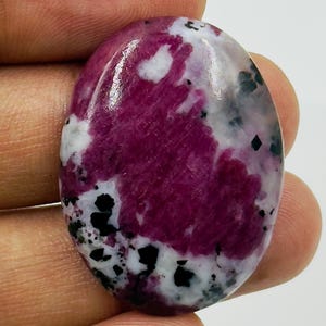 May include: An oval-shaped cabochon gemstone featuring a polished surface with a vibrant magenta-pink centre, contrasted by patches of opaque white and scattered small black inclusions throughout the stone's smooth, rounded surface.