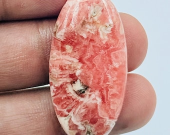 Puede incluir: Un cabujón de piedra preciosa rodocrosita de forma ovalada con una superficie pulida que presenta patrones arremolinados de color rosa salmón, coral e inclusiones minerales de color blanco cremoso en toda la cara.