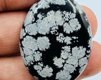 Puede incluir: Una piedra preciosa de obsidiana copo de nieve de forma ovalada con una superficie pulida y lisa. La piedra muestra un color base negro profundo con patrones distintos de color gris claro en forma de flor dispersos por el frente.