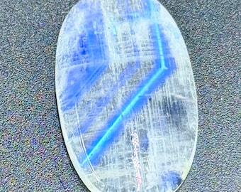 Puede incluir: Un cabujón de piedra lunar de forma ovalada con una base blanca lechosa translúcida y una adularescencia azul vibrante y distintiva que forma una línea diagonal nítida en la superficie, complementada con sutiles inclusiones internas.