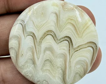 Puede incluir: Un cabujón de piedra pulida de forma circular que presenta una superficie lisa y brillante con patrones intrincados, ondulados y arremolinados en tonos blanco crema, beige y marrón claro, creando un aspecto jaspeado en toda la pieza.