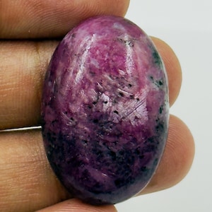 May include: An oval-shaped cabochon gemstone featuring a polished surface with a mix of deep magenta, vibrant pink, and dark black inclusions. The stone displays a smooth, rounded top and a mottled, natural texture throughout its surface.