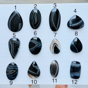 May include: Twelve polished cabochon gemstones arranged in a grid, numbered one through twelve. These stones feature black, white, and grey banded agate patterns in various shapes including teardrop, oval, and rectangular forms with smooth surfaces.