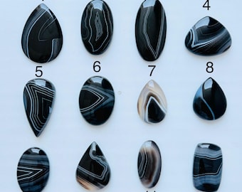 May include: Twelve polished cabochon gemstones arranged in a grid, numbered one through twelve. These stones feature black, white, and grey banded agate patterns in various shapes including teardrop, oval, and rectangular forms with smooth surfaces.