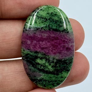 Könnte beinhalten: Ein oval geschliffener Edelstein mit einer lebendigen Farbpalette aus tiefem Magenta und Waldgrün mit schwarzen, gesprenkelten Einschlüssen. Der Stein hat eine glatte, reflektierende Oberfläche und ein deutliches Streifenmuster.