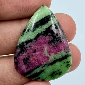 Può includere: Un cabochon di pietra preziosa zoisite lucido a forma di goccia, caratterizzato da un mix vibrante di magenta intenso, verde lime brillante e inclusioni minerali nere scure su tutta la superficie liscia e riflettente della pietra.