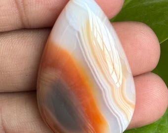 May include: A teardrop-shaped gemstone with bands of white, orange, and gray. The polished stone is held in a hand, with green leaves in the background. The gemstone has a smooth, glossy surface and is likely a cabochon.