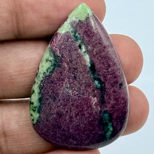 Peut inclure: Une pierre précieuse polie en forme de goutte d'eau présentant une couleur de base magenta et violet profond avec des stries et des taches proéminentes d'inclusions minérales vert clair et vert forêt foncé sur toute la surface lisse et brillante.