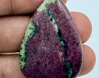 Puede incluir: Una piedra preciosa pulida en forma de lágrima que presenta un color base magenta y púrpura intenso con vetas y parches prominentes de inclusiones minerales de color verde claro y verde bosque oscuro en toda la superficie lisa y brillante.