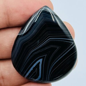 May include: A polished, teardrop-shaped black agate gemstone featuring distinct, sharp white and light gray concentric banding patterns that follow the contour of the stone, creating a high-contrast, layered appearance across the smooth surface.