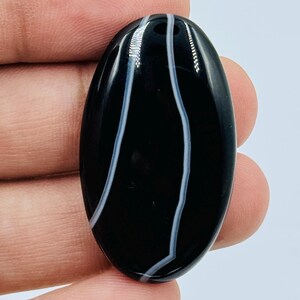 May include: An oval-shaped black gemstone cabochon featuring two distinct, vertical, wavy white bands running through the center. The polished stone has a smooth, reflective surface and a dark, opaque appearance with high-contrast white lines.