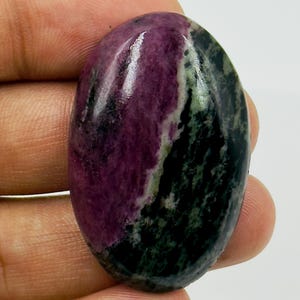 Peut inclure: Une pierre précieuse polie de forme ovale présentant une séparation de couleurs distincte avec une section magenta-pourpre profond sur la gauche et une section vert forêt foncé et noire tachetée sur la droite, à la surface lisse et réfléchissante.