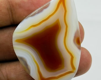 Puede incluir: Una piedra preciosa de ágata pulida en forma de lágrima que presenta bandas concéntricas de blanco translúcido, naranja vibrante y marrón rojizo intenso. La piedra tiene una superficie lisa y brillante con un patrón en capas definido.