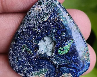 Puede incluir: Una piedra de azurita triangular, predominantemente azul oscuro con inclusiones verdes y blancas. La piedra pulida muestra patrones naturales y se sostiene en una mano. La piedra mide aproximadamente 5 cm.